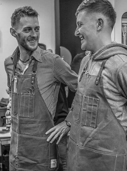 Two of the barbers in canvas aprons, sharing a laugh in the shop